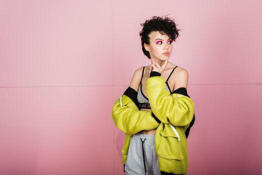 Beautiful teenager girl sincerely smiles and laughs in sports stylish clothes in the studio on a pink background. Bright girl with a short flying hairstyle in a puffed neon lemon jacket and pink smokey eyes.