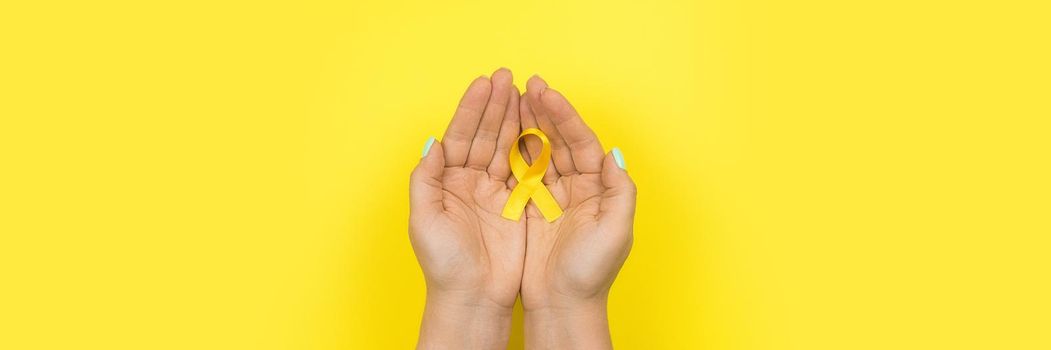 Yellow gold ribbon in the hands of a girl on a yellow background. Children cancer concept. Awareness month. Childhood Cancer Day February, 15. Web banner.