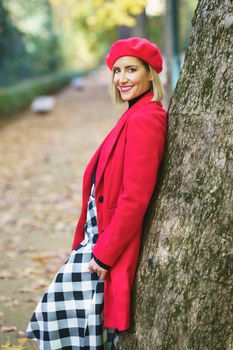 Side view of smiling female wearing red outerwear and checkered shirt leaning on tree in autumn park and smiling at camera