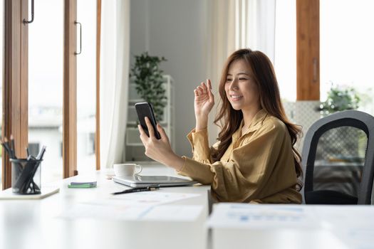 Happy young business asian woman holding phone looking at screen waving hand video calling distance friend online in mobile chat app using smartphone application