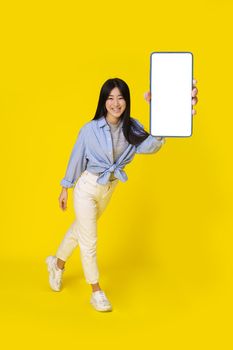 Charming young asian girl holding smartphone showing white screen mobile app advertisement and excited smile on camera wearing casual isolated on yellow background. Product placement.