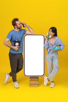 Love talks, handsome guy and asian girl using vintage phones leaned on huge smartphone on old books with blank screen, happy smiling isolated on yellow background. Mock up, product placement.