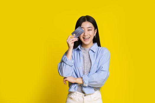Happy excited young asian girl with credit, debit card. Asian girl in blue shirt holding mockup credit card isolated on yellow background. E-banking concept. E-commerce concept.