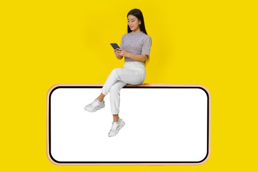 Asian girl with phone in hands sit on huge smartphone with white screen wearing casual white jeans isolated on yellow background. Mock up product placement mobile app advertisement. Copy space.