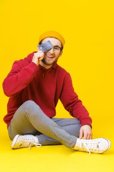 Happy young hipster man in red hoodie sitting on the floor holding with credit, debit card buying, selling, money transfer concept. Freelancer caucasian man isolated on yellow background.
