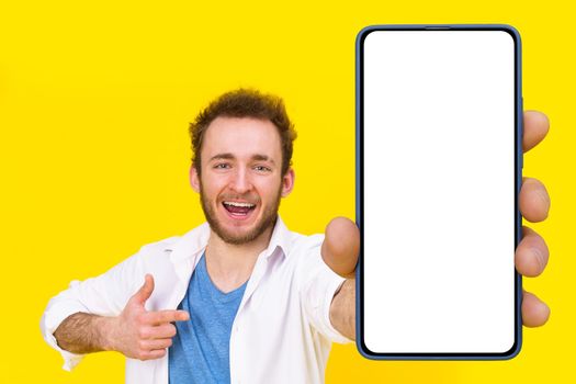 Check this app young man pointing at smartphone showing a white empty screen game, bet, lottery win isolated over yellow background. Product placement for mobile application advertisement.
