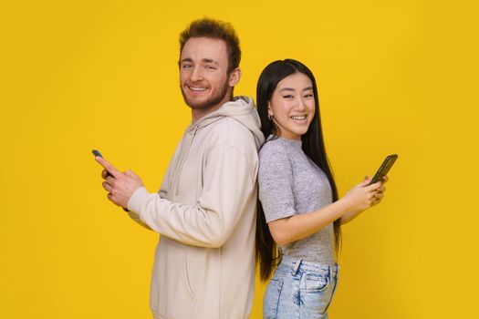 Caucasian guy and asian girl standing back to back smiling holding smartphones in hand looking at camera mobile app advertisement isolated on yellow background. Product placement.