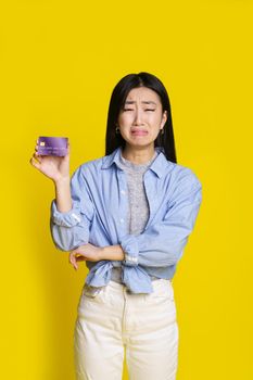 Crying asian girl holding purple credit, debit card with sad, unhappy face begging for money. Asian girl in blue shirt with mockup credit card isolated on yellow background. Shopping concept.