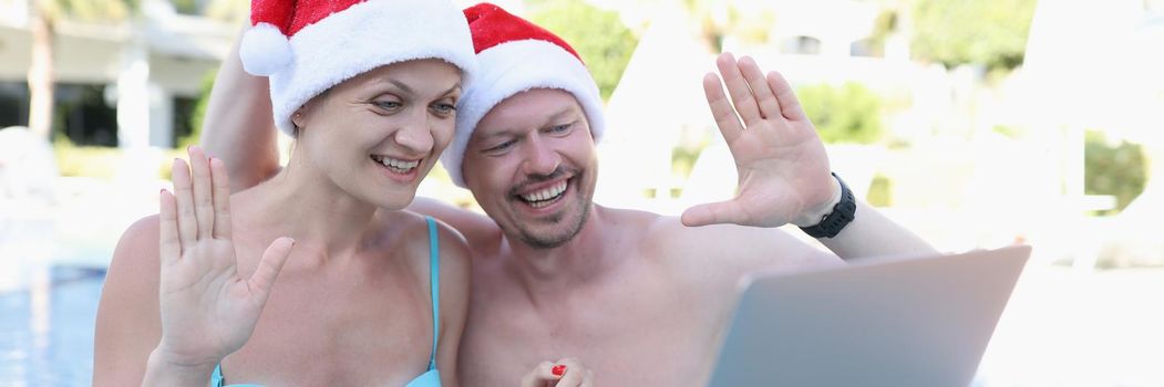 A man and a woman in santa claus hats in the pool use a laptop, close-up. Video conference, remote access