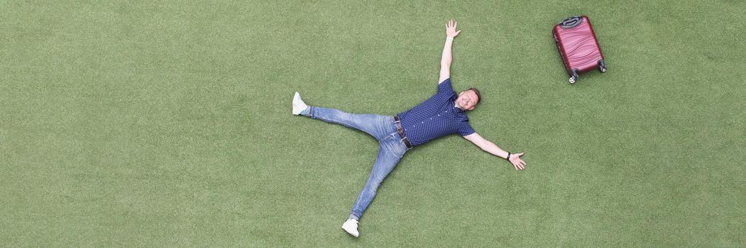 Happy man with a suitcase lies on a green lawn, top view. Arrival at the resort, vacation alone. Freedom from work