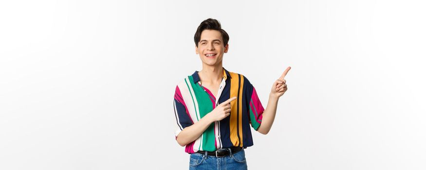 Carefree guy in stylish shirt pointing fingers at upper right corner, showing promo banner or logo, smiling and talking to someone, standing over white background.