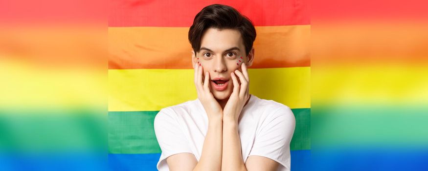 Close-up of beautiful brunette gay man in white t-shirt, with glitter makeup, looking amused at camera, touching face, standing against rainbow pride flag.