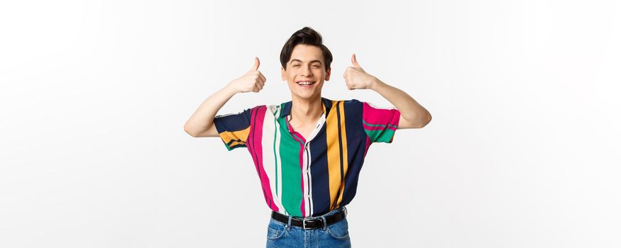 Young cheerful man smiling, showing thumbs up in approval, praise something good, standing over white background.