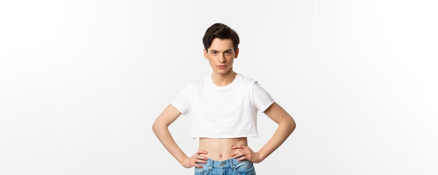 Handsome androgynous man in crop top looking confident at camera, holding hands on waist. Lgbtq activist standing over white background with sassy expression.