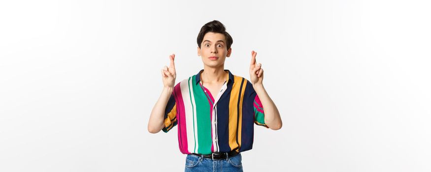 Hopeful young androgynous man cross fingers for good luck, making wish, waiting for something, standing over white background.