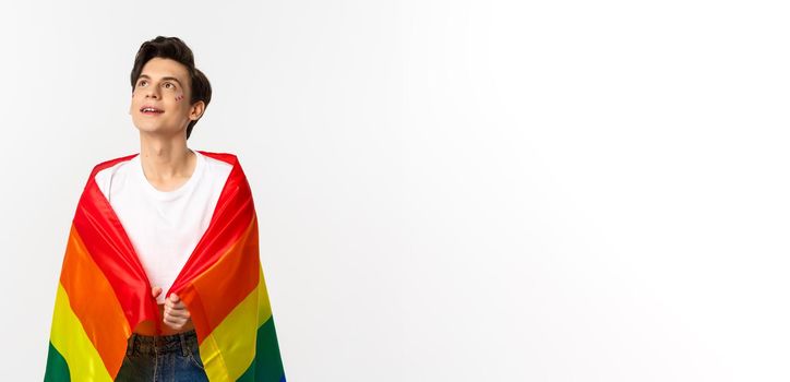 Human rights and lgbtq community concept. Beautiful young androgynous man with glitter on face, wearing rainbow pride flag and looking at upper left corner, white background.