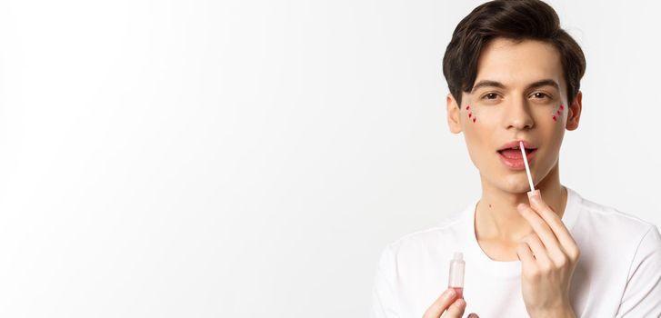 Close-up of gender fluid person with glitter on face, applying lip gloss lipstick and looking at camera, standing against white background.