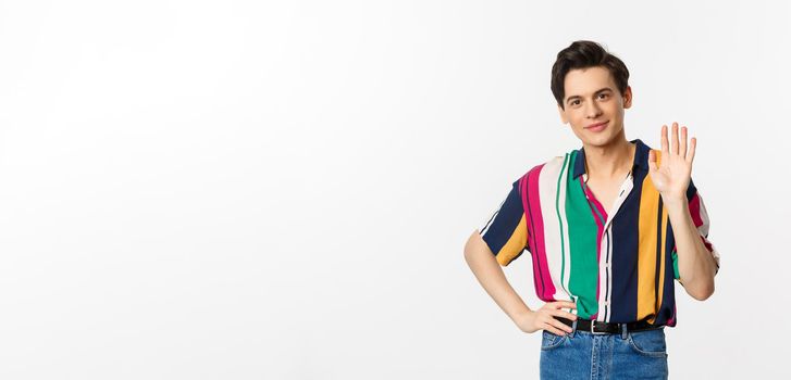 Friendly young man smiling and saying hello, waving hand in greeting gesture, standing over white background.