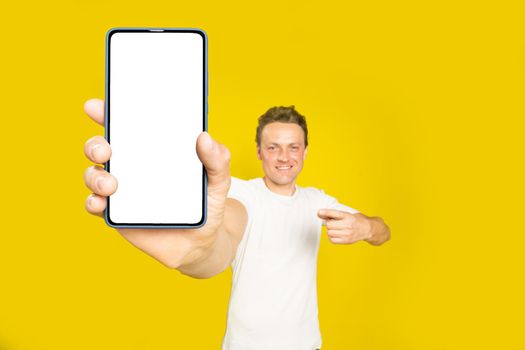 Mobile app advertising handsome blond man pointing at giant smartphone with white empty screen in his hand, wearing white t-shirt and jeans isolated on yellow background. Great offer.