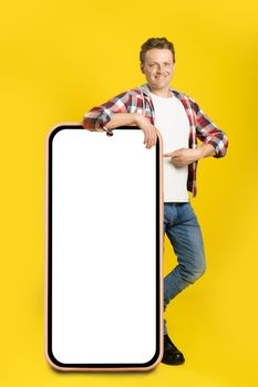 Pointing finger handsome caucasian man leaned on giant, huge smartphone with blank white screen, wearing casual outfit isolated on yellow background. Free space mock up.