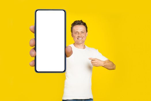 Handsome blond man pointing at giant smartphone with white empty screen, wearing white t-shirt and jeans cellphone display mock up isolated on yellow background. Mobile app advertisement.