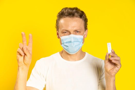 Happy man gesturing victory holding test wearing medical face mask pandemic coronavirus or monkeypox. Handsome man in white t-shirt and medical mask on yellow background. Healthcare concept.