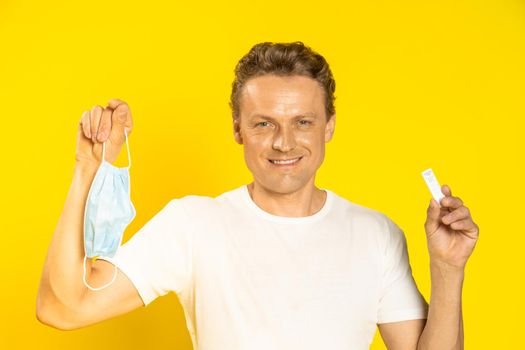 Happy man holding test and medical face mask in hands celebrating victory pandemic coronavirus or monkeypox. Handsome man in white t-shirt and medical mask on yellow background. Healthcare concept.