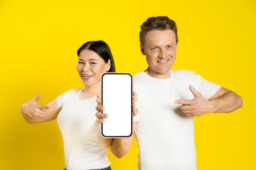 Caucasian man and asian woman show thumbs up and pointing finger excited holding smartphone with blank white screen, mobile app advertisement isolated on yellow background. Product placement.