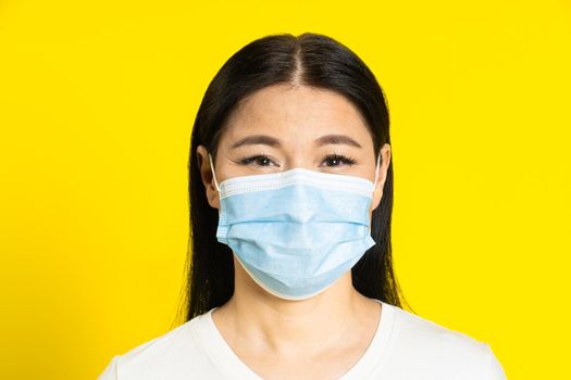 Happy, safe of pandemic mature asian woman wearing medical face mask coronavirus or monkeypox prevention. Charming middle age woman in white t-shirt and medical mask on yellow background.