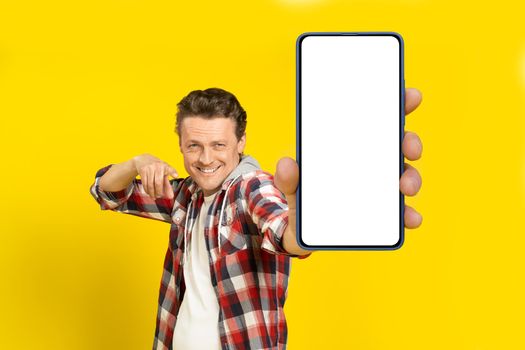 Yo, look at this. Happy handsome man pointing at huge smartphone with white empty screen, wearing red plaid shirt, cellphone display mock up isolated on yellow background. Mobile app advertisement.