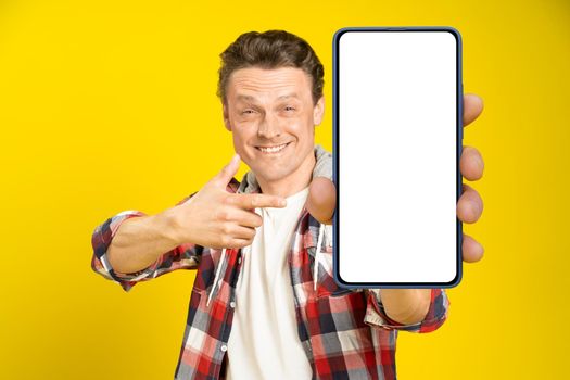 Charming happy man pointing at smartphone with white empty screen, wearing red plaid shirt and jeans cellphone display mock up isolated on yellow background. Mobile app advertisement.