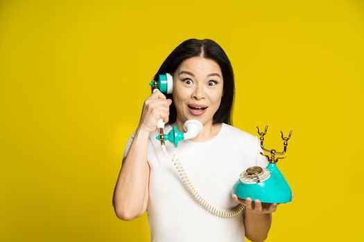 Funny face expression asian woman using vintage, retro telephone in hands wearing white t-shirt isolated on yellow background. Excited asian woman with retro phone. Communication concept.