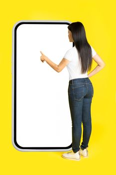 Charming middle aged asian woman with phone in hands showing white screen leaned back on giant, huge smartphone with white screen wearing casual isolated on yellow background. Free space mock up.