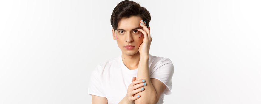 People, lgbtq and beauty concept. Close-up of beautiful queer man touching face with fingers with blue nail polish, standing over white background.