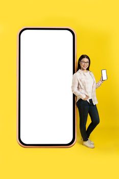 Charming middle aged asian woman with phone in hands showing white screen leaned back on giant, huge smartphone with white screen wearing casual isolated on yellow background. Free space mock up.
