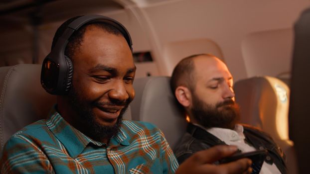 African american tourist using smartphone to have fun on flight, travelling by airplane in economy class. Browsing internet on mobile phone and flying on plane during sunset.