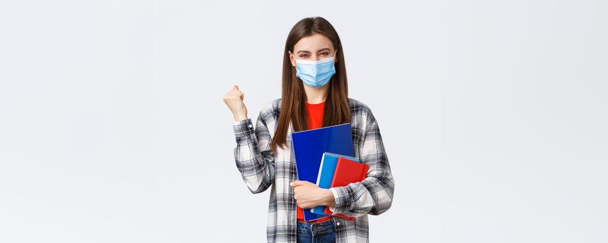 Coronavirus pandemic, covid-19 education, and back to school concept. Rejoicing, cheerful female student in medical mask happy entering cool university, fist pump in success, holding notebooks.