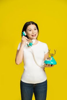 Charming funny asian woman using vintage, retro telephone in hands wearing white t-shirt isolated on yellow background. Excited asian woman with retro phone. Communication concept.