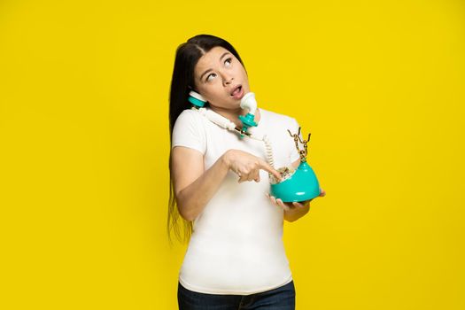 Funny asian woman using vintage, retro telephone in hands dreamingly dialling trying to remember phone number face expression wearing white t-shirt isolated on yellow background.