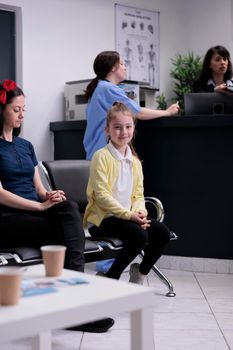 Portrait of smiling young girl with her mother sitting in hospital lobby waiting to see pediatrician doctor at private clinic.