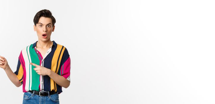 Amazed young queer man pointing fingers left, staring at camera with disbelief, asking question about product, standing over white background.