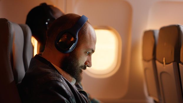 Businessman feeling tired and angry about work trip on plane, browsing internet on laptop during sunset. Flying in economy class with passengers, working on computer and travelling.