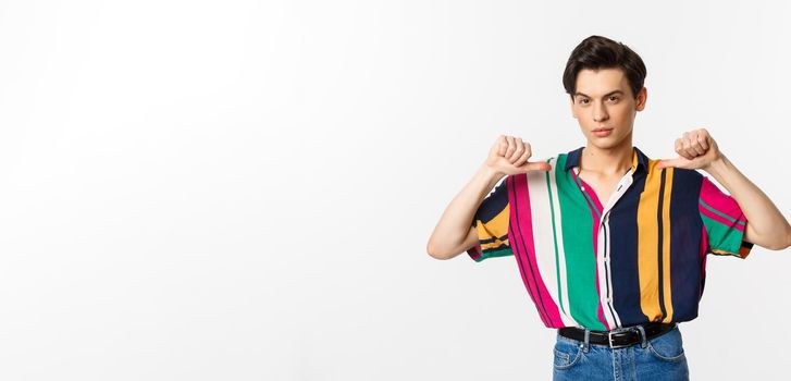 Confident and sassy gay man pointing at himself, looking self-assured, standing over white background.