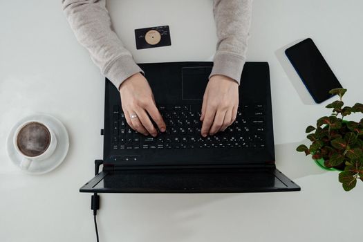 woman working on laptop at home. Coffee, phone, houseplant, credit card on the white table. Wonam typing on laptop