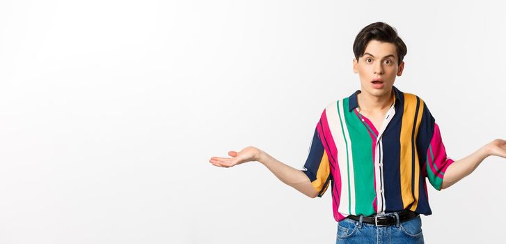 Confused young androgynous man spread hands and looking clueless, dont understand, standing puzzled against white background.