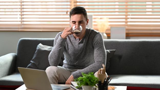 Young man drinking coffee and using computer laptop on couch.