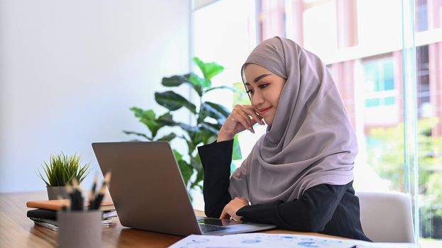 Beautiful young Muslim business woman in hijab sitting at modern office and watching online webinar on laptop.