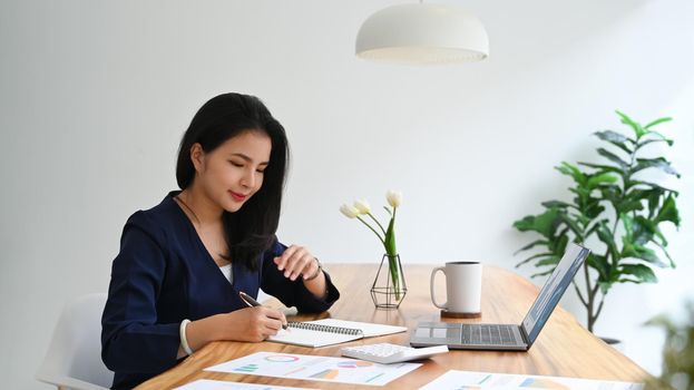 Happy young woman working with financial document and using laptop on wooden office desk.