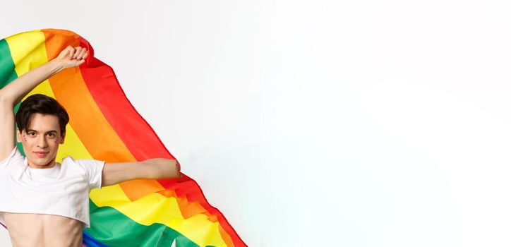 Vertical view of happy queer person in crop top and jeans waving raised rainbow flag, celebrating lgbtq holiday, standing over white background.