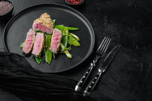 Homemade Cut Grilled Sesame Tuna steak set with spring onions and sugar snap peas, on plate, on black stone background , with copyspace and space for text